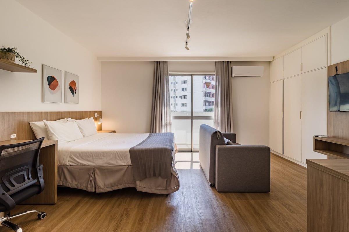 Quarto de hotel com cama, escrivaninha, cadeira, guarda-roupa e janela com cortinas.