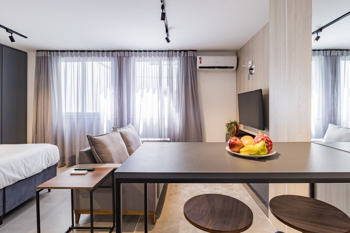 Ett modernt hotellrum med en säng, ett skrivbord, ett matbord med frukt, en tv och ett fönster med gardiner.