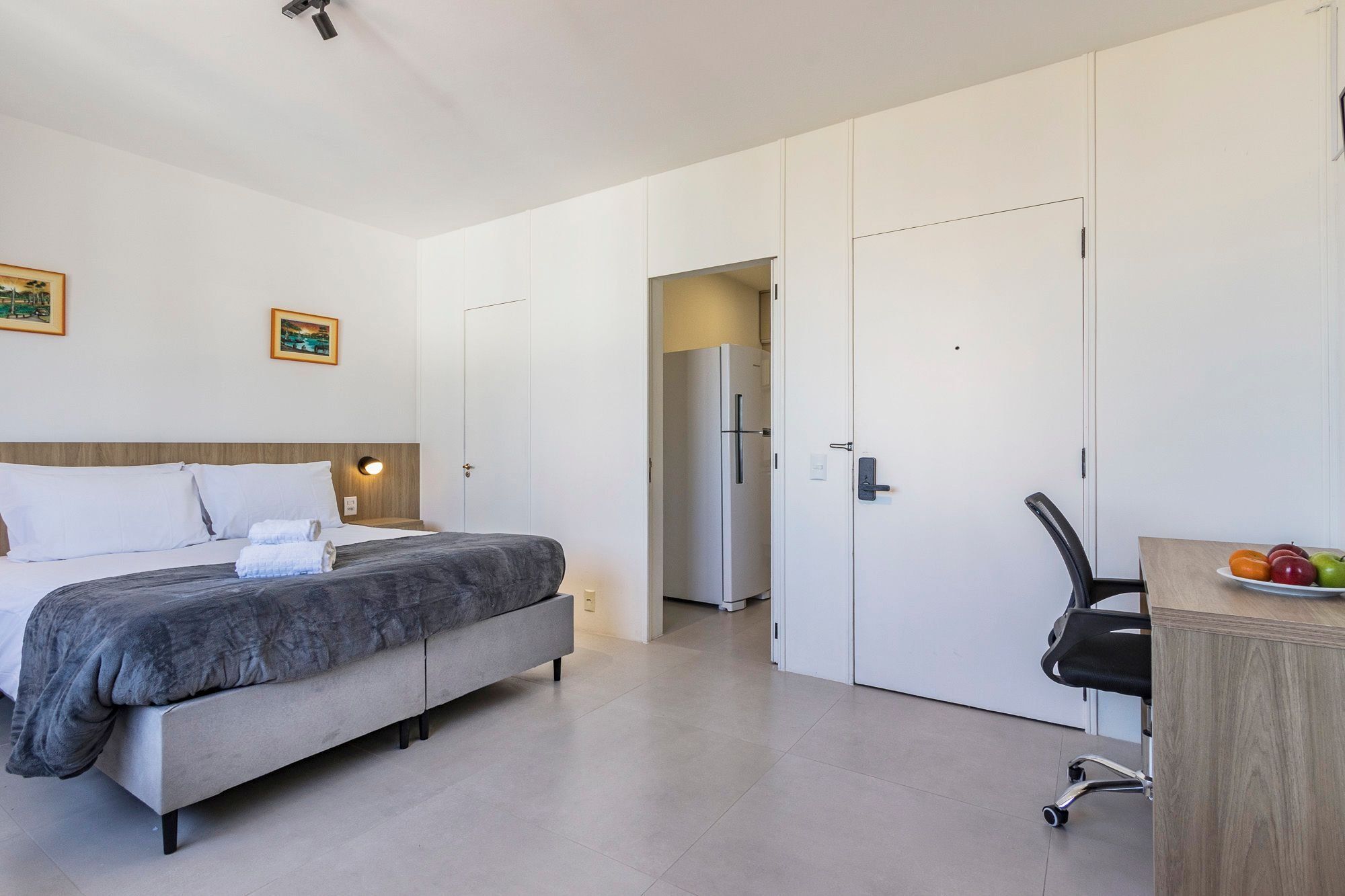 A hotel room with a bed, wardrobe, desk, and a small dining area with fruit.