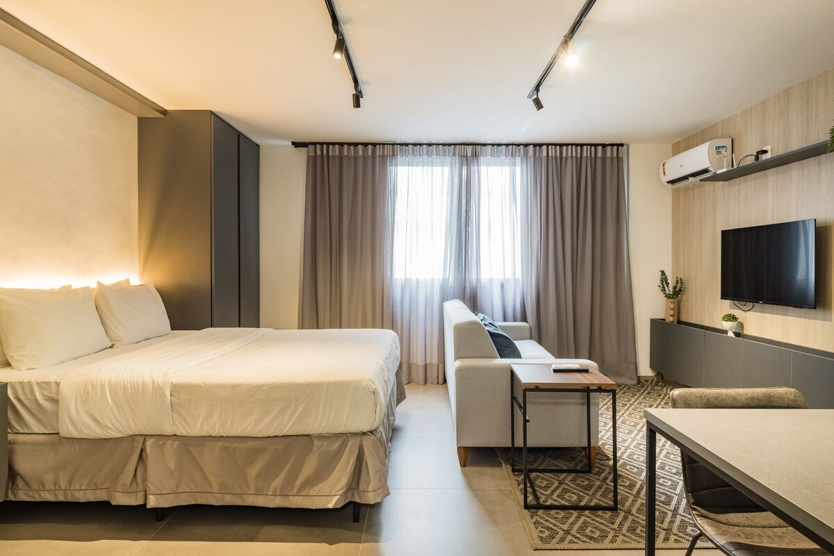 A hotel room with a bed, a sofa, a desk, a TV, and an air conditioning unit.