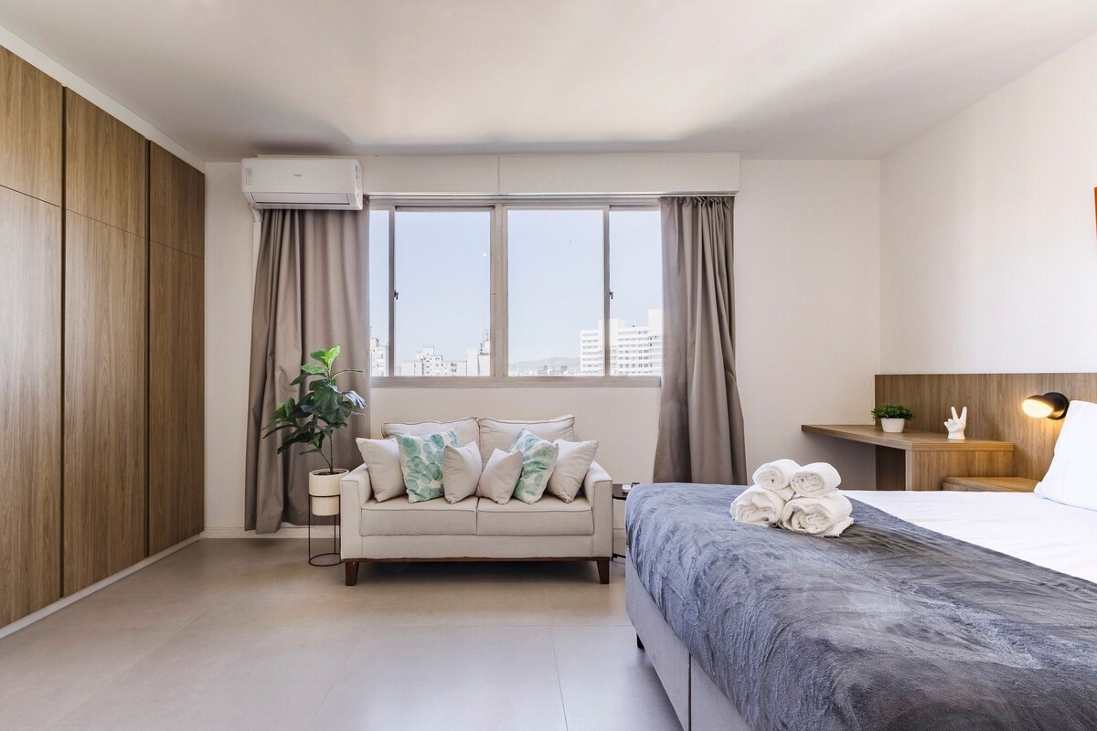 A modern hotel room with a sofa, a bed, a potted plant, and a view of the city.