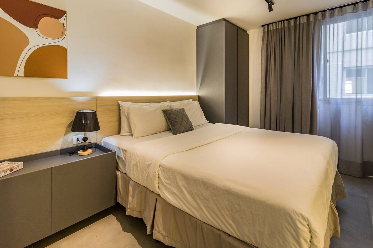 Quarto de hotel com cama, mesa de cabeceira, abajur e cortinas.