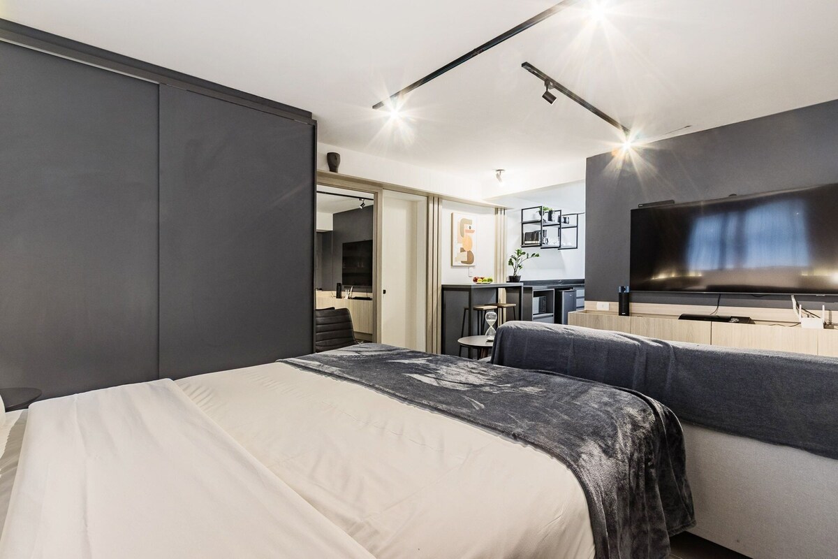 A modern hotel room with a bed, a TV, a desk, and a kitchenette.