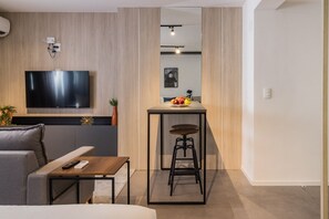 A modern living room with a flat-screen TV, a small table with a remote, a sofa, and a kitchen area with a bar stool and a fruit bowl. - Duque by My Way (Porto Alegre)