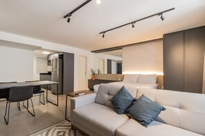 A modern living room with a sofa, a dining table with chairs, a kitchen area, and a large mirror. - Duque by My Way (Porto Alegre)