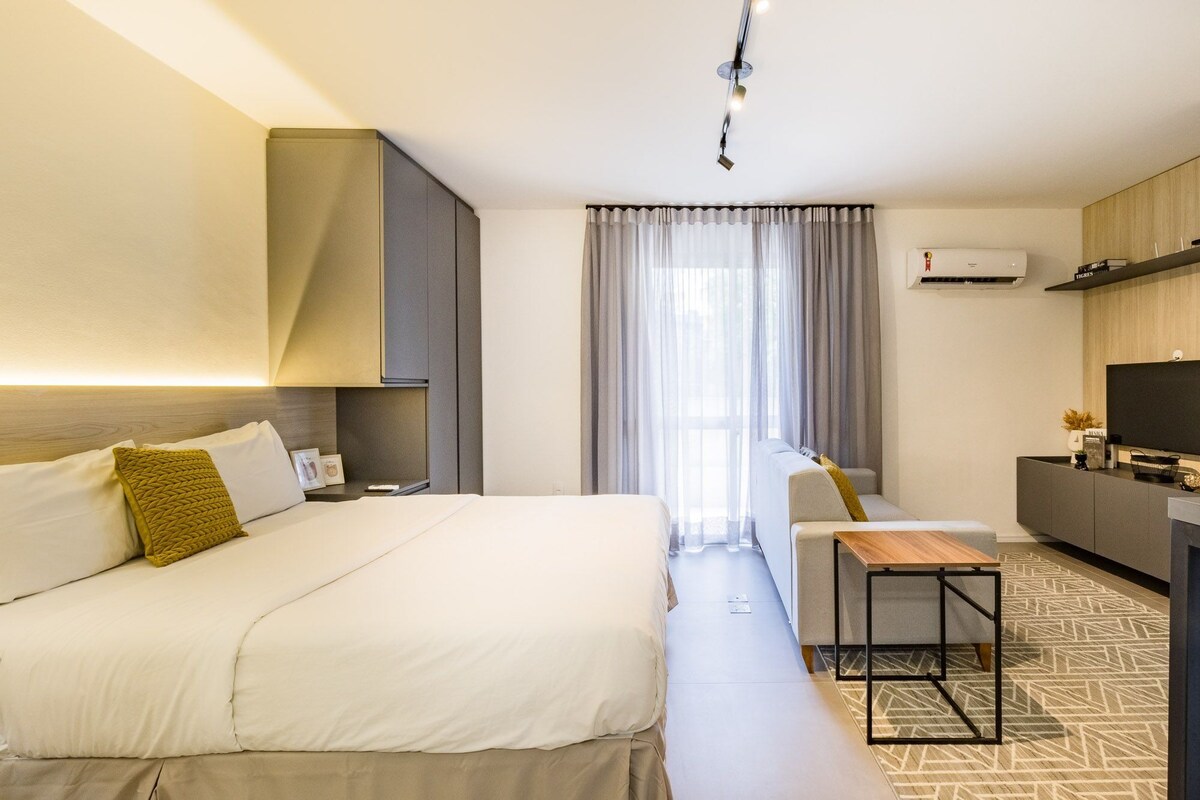A hotel room with a bed, a sofa, a small table, a TV, and an air conditioning unit.