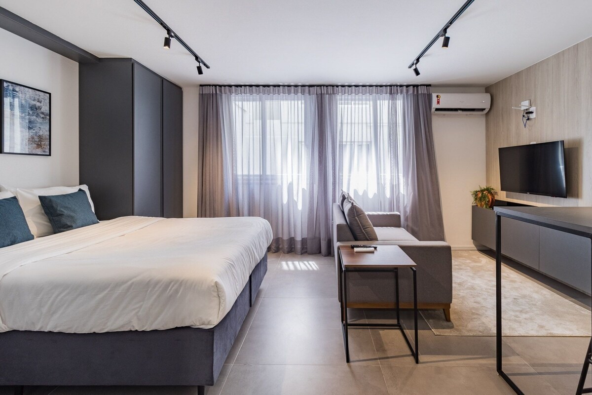 A modern hotel room with a bed, a sofa, a TV, and an air conditioning unit.