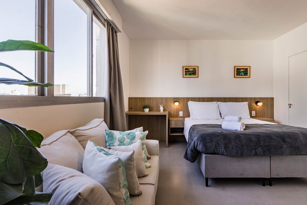 Une chambre d’hôtel comprenant un lit, un canapé, une table de chevet, une fenêtre avec des rideaux et deux tableaux accrochés au mur.