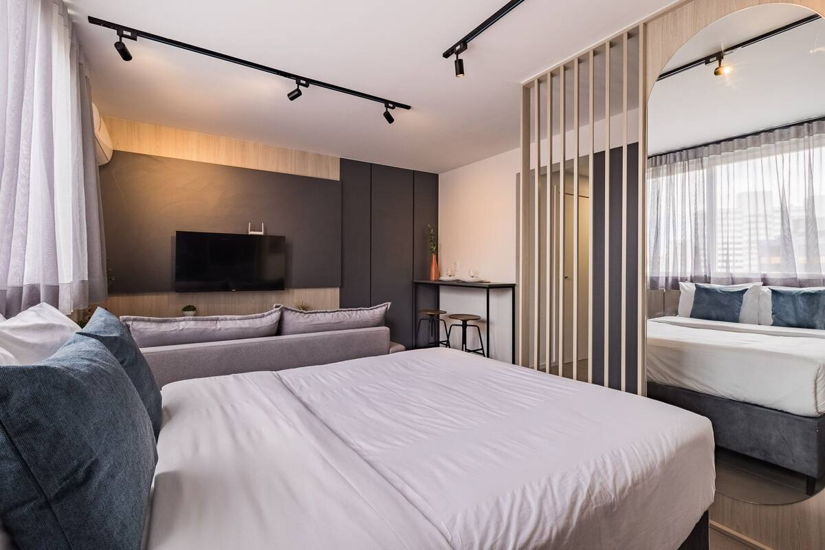 Quarto de hotel moderno com uma cama grande, um sofá, uma TV fixada na parede, uma mesa pequena com banquetas e uma janela com cortinas leves.