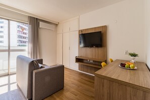 A modern hotel room with a sofa, a dining table with fruit, a TV, and a wardrobe. - Duque by My Way (Porto Alegre)
