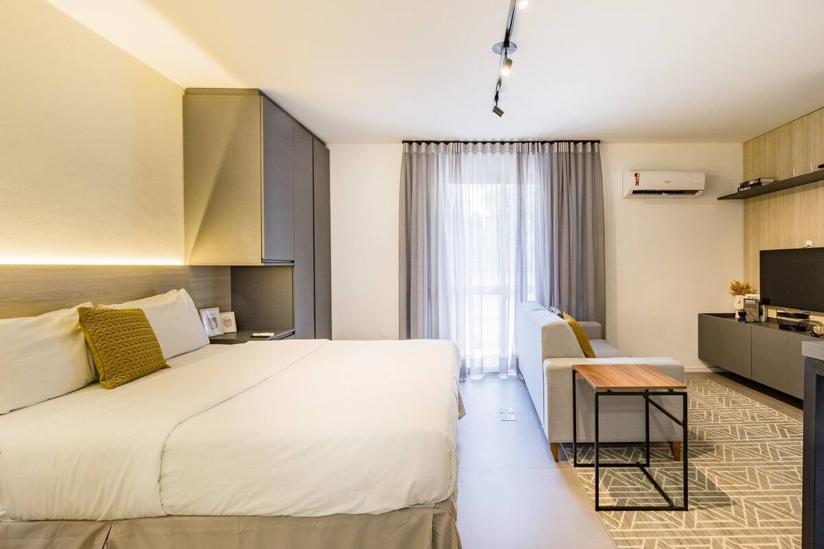 Quarto de hotel com cama, sofá, mesinha, televisão e ar condicionado.