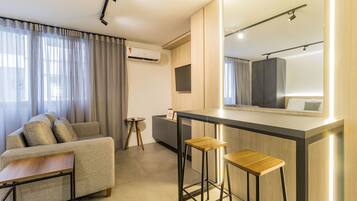 A modern hotel room with a sofa, a small round table, a bar area with stools, a large mirror, and an air conditioning unit.