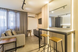 A modern hotel room with a sofa, a small round table, a bar area with stools, a large mirror, and an air conditioning unit. - Duque by My Way (Porto Alegre)