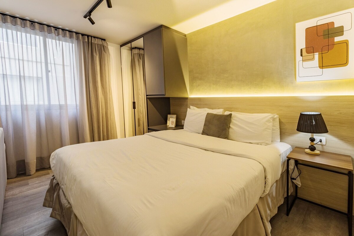 A hotel room with a large bed, a nightstand with a lamp, and a wall-mounted artwork.