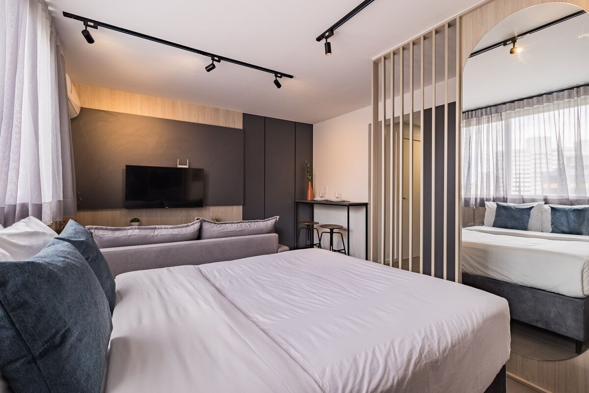 A modern hotel room with a large bed, a sofa, a TV mounted on the wall, a small table with stools, and a window with sheer curtains.