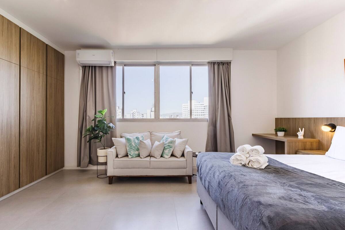 Quarto de hotel moderno com sofá, cama, planta em vaso e vista da cidade.