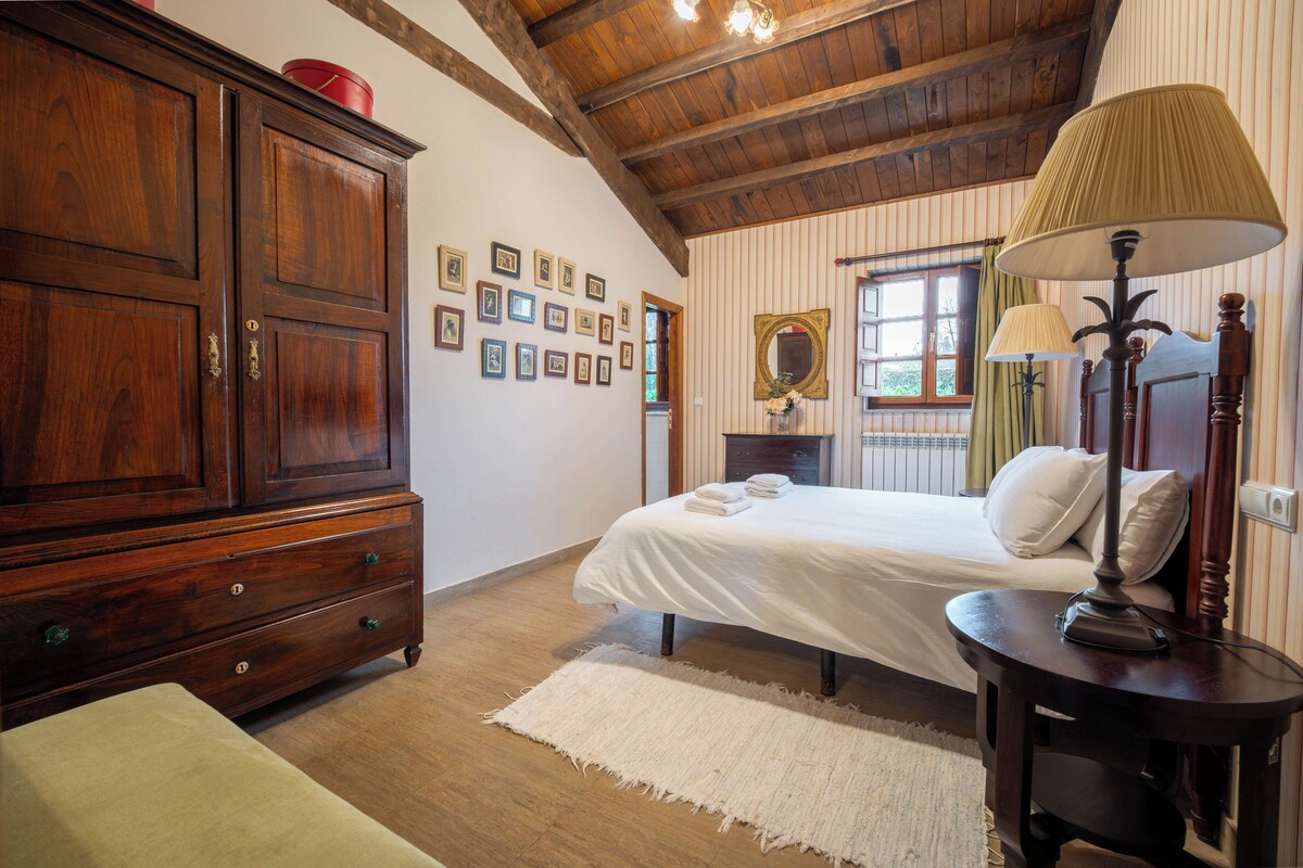 A bedroom with a wooden wardrobe, a bed with white linens, a bedside table with lamps, and a large mirror on the wall.