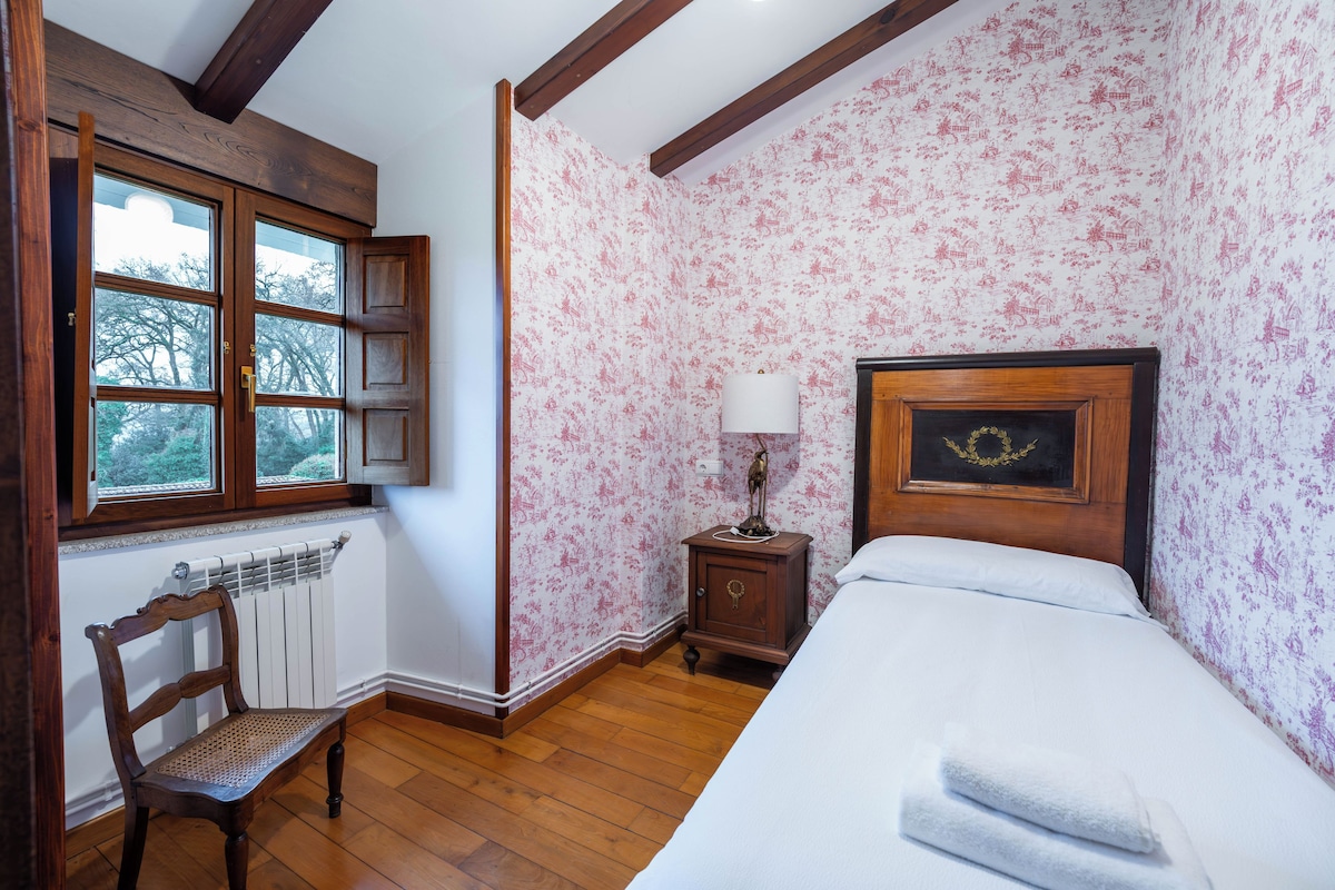 A bedroom with a wooden bed, a nightstand with a lamp, a chair, a window with a view, and floral wallpaper.