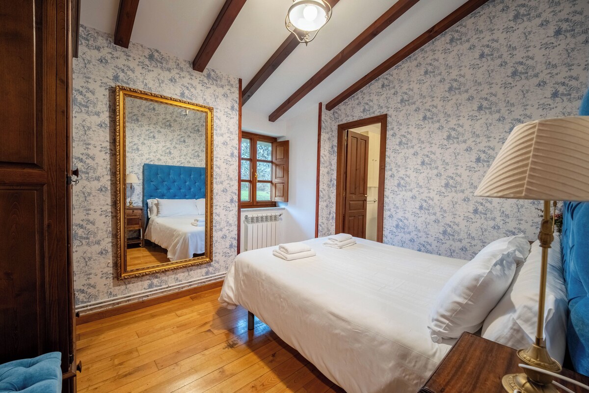 A bedroom with a large mirror, a bed with white linens, a wooden floor, and a window with a view of greenery.