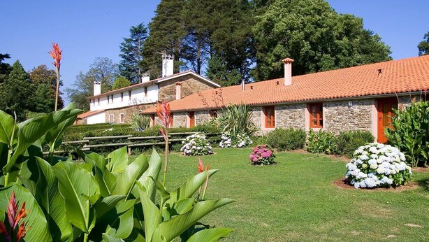 A stone building with a red-tiled roof, surrounded by a well-maintained lawn and various plants. - A Painza Aldea Rural  (Arzua)