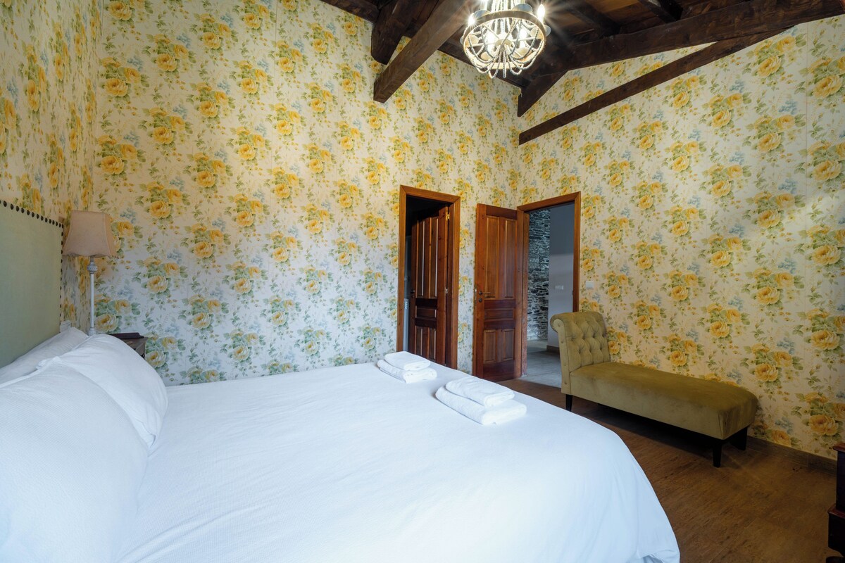 A room with floral wallpaper, a large bed with white linens, a bench, and a chandelier.