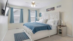 Un dormitorio con cama, mesita de noche, ventilador de techo y ventana con cortinas.