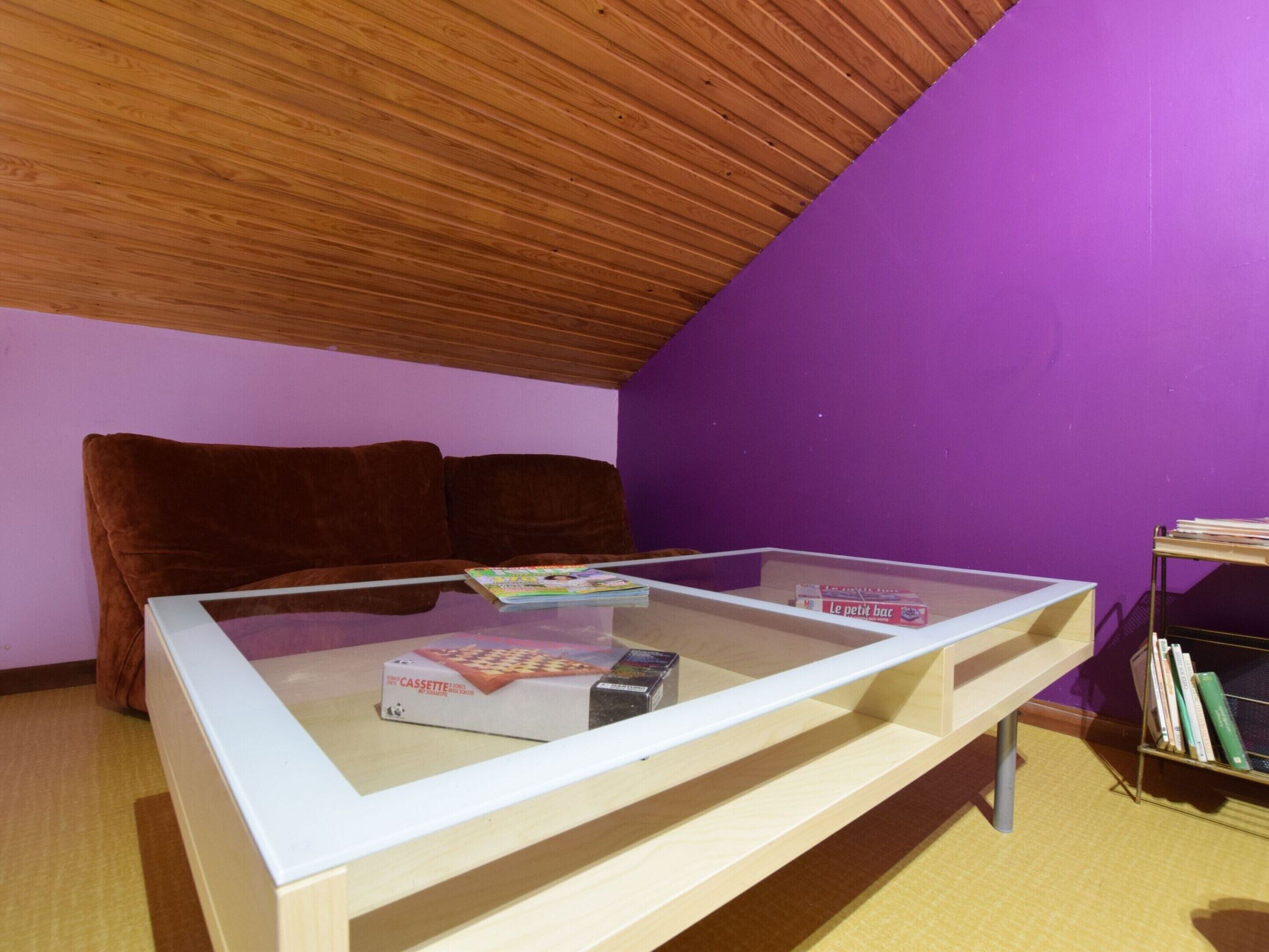 Una habitación con techo inclinado de madera, pared morada, sofá marrón, una mesa de vidrio con libros y un reproductor de casetes, y una estantería con libros.
