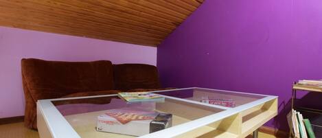 Una habitación con techo inclinado de madera, pared morada, sofá marrón, una mesa de vidrio con libros y un reproductor de casetes, y una estantería con libros.