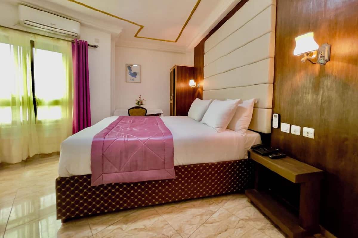 Quarto de hotel com cama grande, mesa de cabeceira, luminária e janela com cortinas.