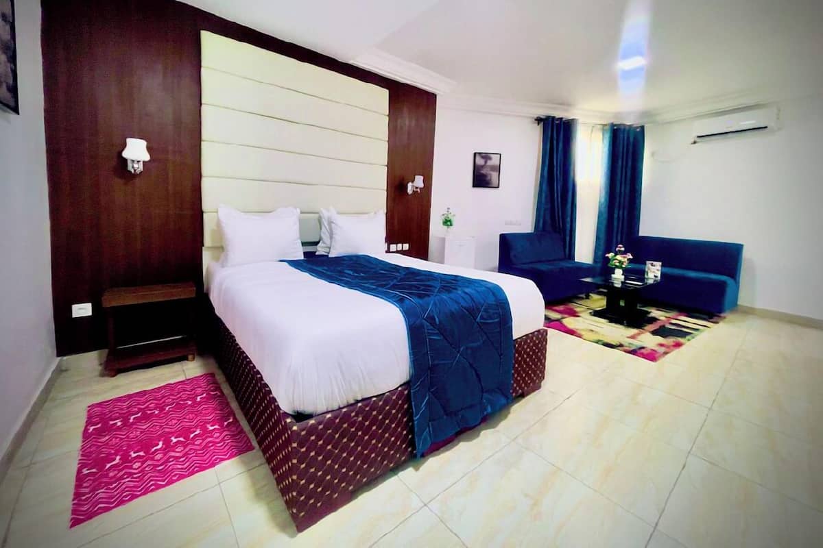 Quarto de hotel com uma cama grande, um sofá azul, uma mesa pequena com flores e um ar condicionado fixado na parede.
