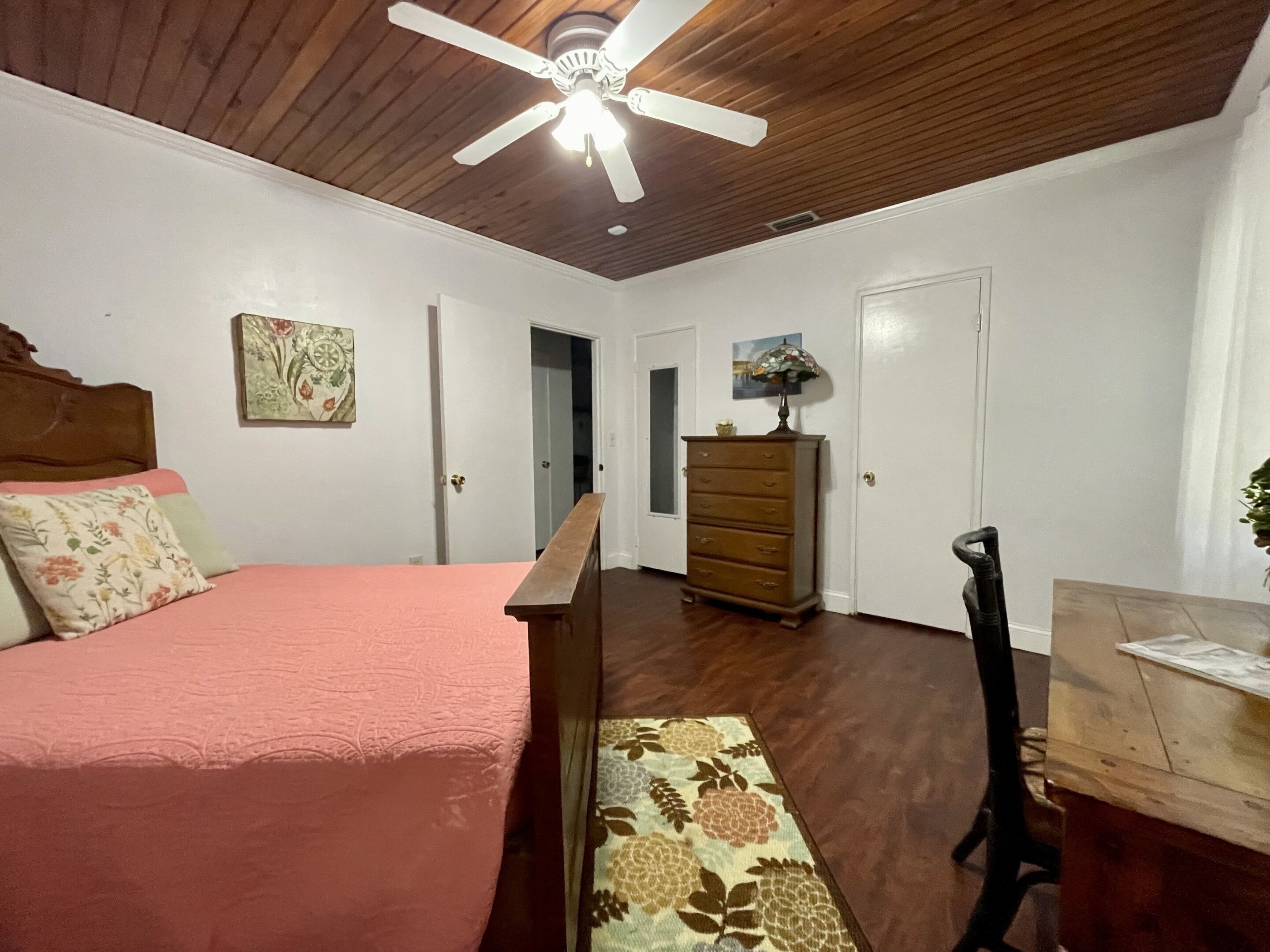 A bedroom with a bed, a dresser, a desk, a ceiling fan, and a door.