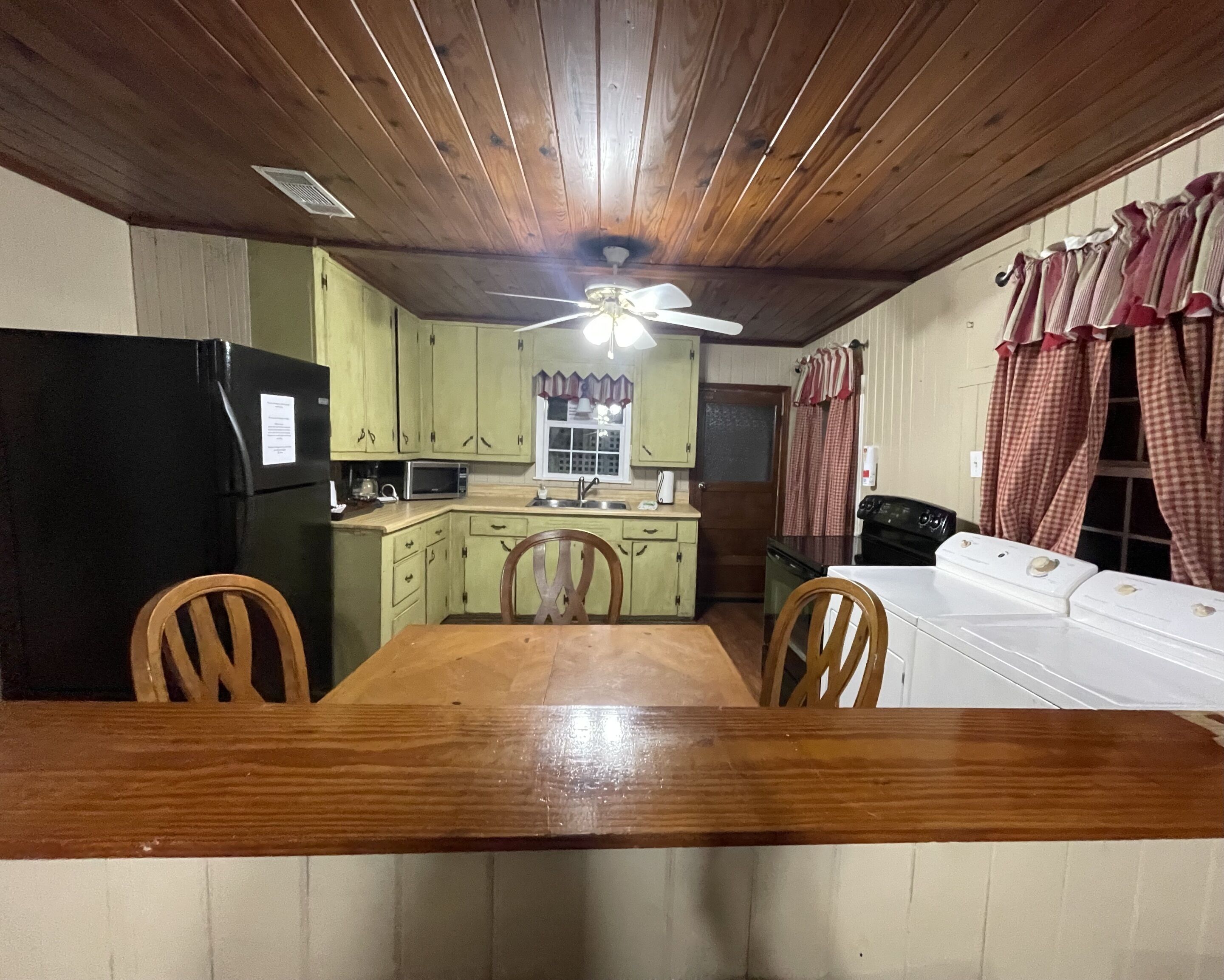 Uma cozinha com armários de madeira, uma mesa de jantar, cadeiras, um ventilador de teto e uma máquina de lavar.