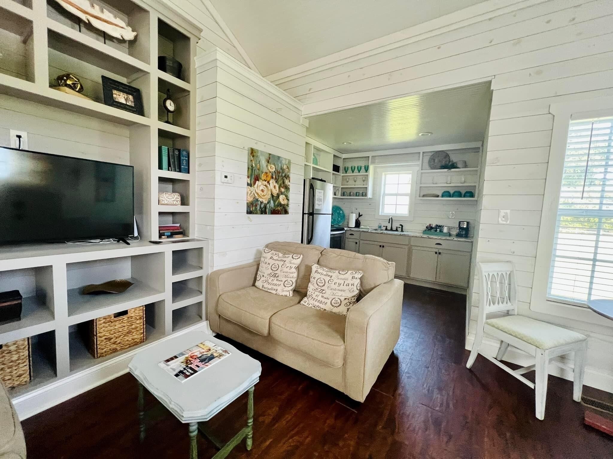 A living room with a beige sofa, a television, and a coffee table. There is a kitchen in the background with white cabinets and a refrigerator.