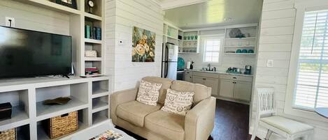 A living room with a beige sofa, a television, and a coffee table. There is a kitchen in the background with white cabinets and a refrigerator.