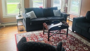 A living room with a black sofa, a wooden coffee table, and a patterned rug. There are three windows with white blinds, a ceiling fan, and a lamp on a side table. - Cozy 1-bedroom apartment with WiFi, AC, full kitchen, in enjoyable Colebrook (Colebrook)