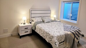 A bedroom with a bed, two bedside tables, a window with blinds, and a nightstand with a lamp.
