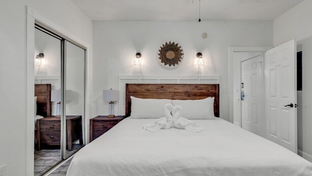 A hotel room with a wooden headboard, a large bed with white linens, two bedside lamps, a wall-mounted decorative item, and a sliding door closet. - Elegant 3BR Condo: Magnolia North 101 (Myrtle Beach)