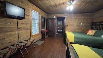 A room with wooden walls, a bed with a green and yellow bedspread, a television on the wall, and a ceiling fan.
