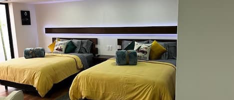Habitación de hotel con dos camas, cada una con una colcha amarilla y varias almohadas. El piso es de madera y el techo tiene un diseño moderno con iluminación empotrada.