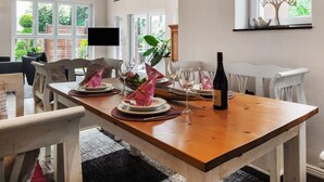 Un comedor con una mesa de madera dispuesta para cuatro personas, rodeada de sillas blancas. Hay una botella de vino sobre la mesa, y un televisor está montado en la pared encima de la mesa.