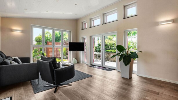 Un salón espacioso con un sofá gris, un sillón negro y un televisor de pantalla plana grande. Varias ventanas ofrecen luz natural y vistas al jardín.