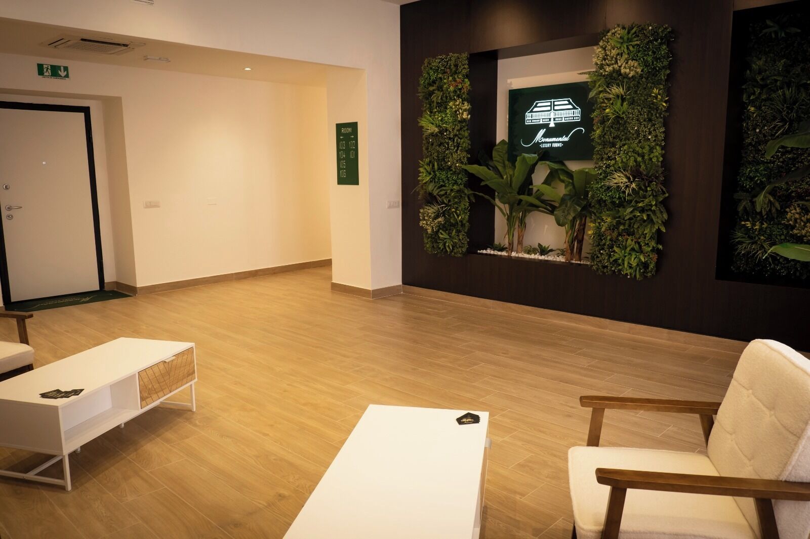A reception area with a wooden floor, a white reception desk, a seating area with a white sofa and a wooden chair, and a wall-mounted display with a green wall and a sign indicating room numbers.