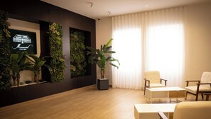 Un área de espera moderna con piso de madera, dos sillas blancas, una planta grande en maceta y una pantalla montada en la pared.