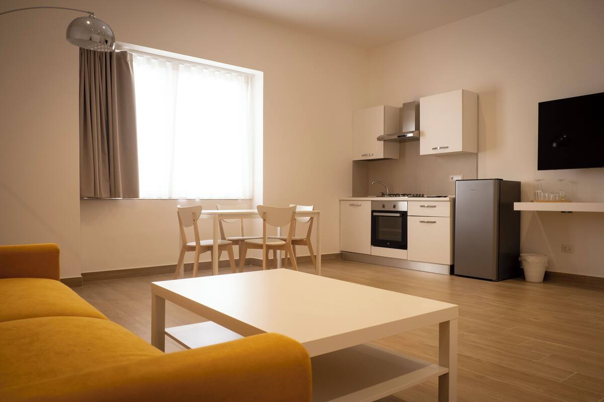 A modern kitchen with white cabinets, a built-in oven, and a refrigerator. There is a dining area with a white table and chairs.