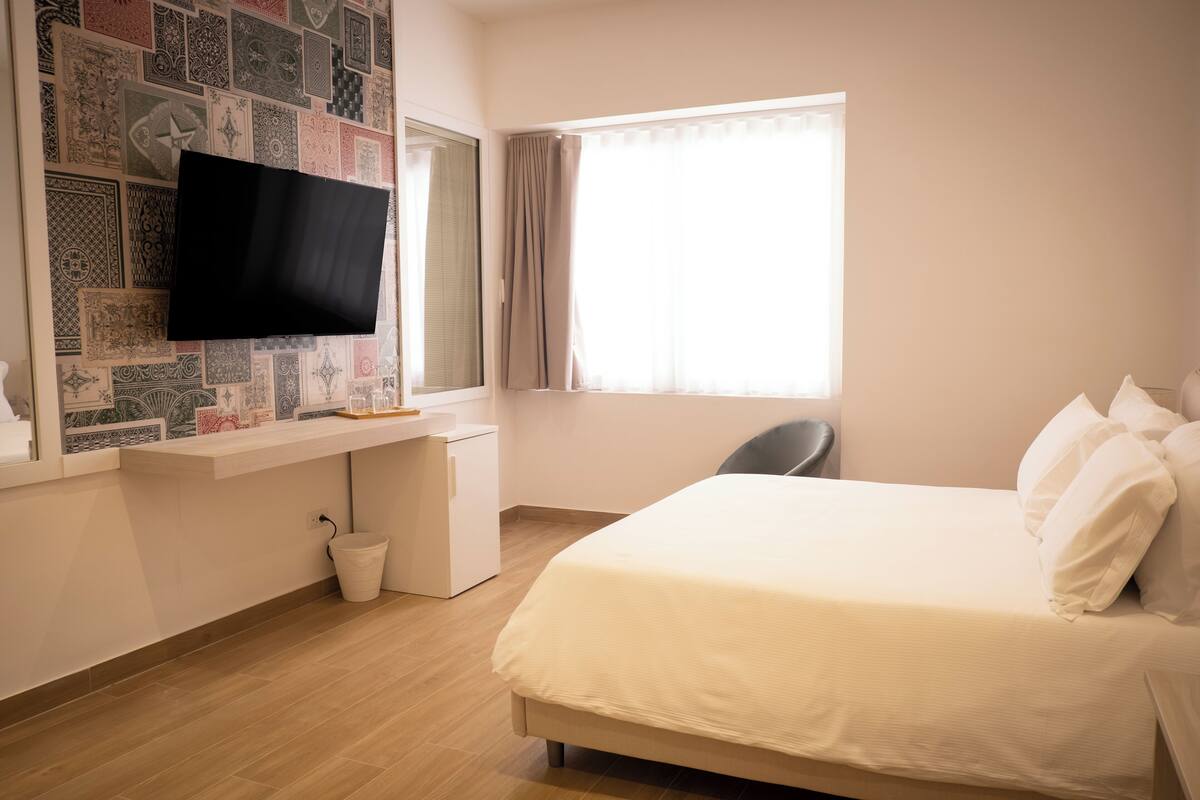 A hotel room with a bed, a desk with a flat-screen TV, a chair, and a window with curtains.