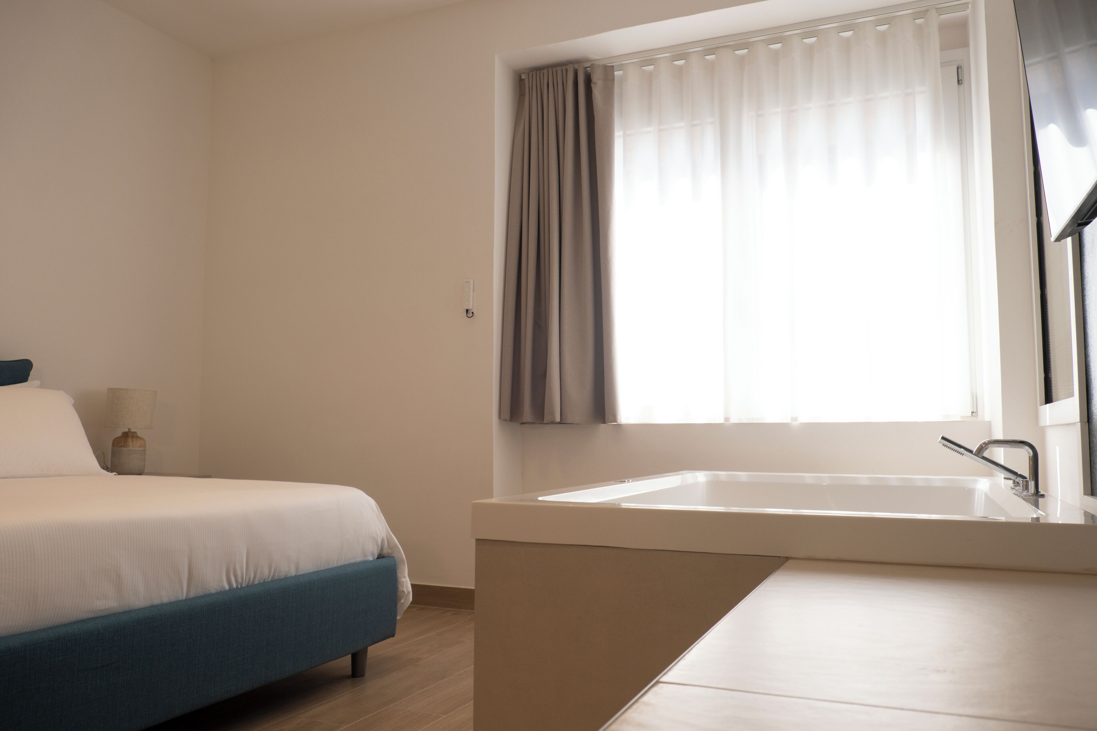 A bedroom with a bed, a nightstand, a window with curtains, and a bathroom with a sink.