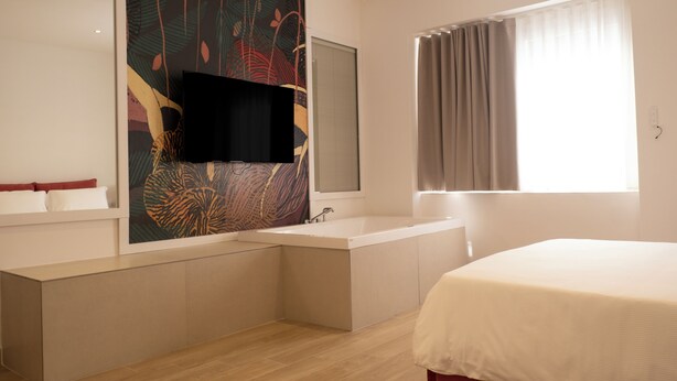 Una habitación de hotel moderna con una cama grande, una bañera integrada, un televisor montado en la pared y una ventana con cortinas.
