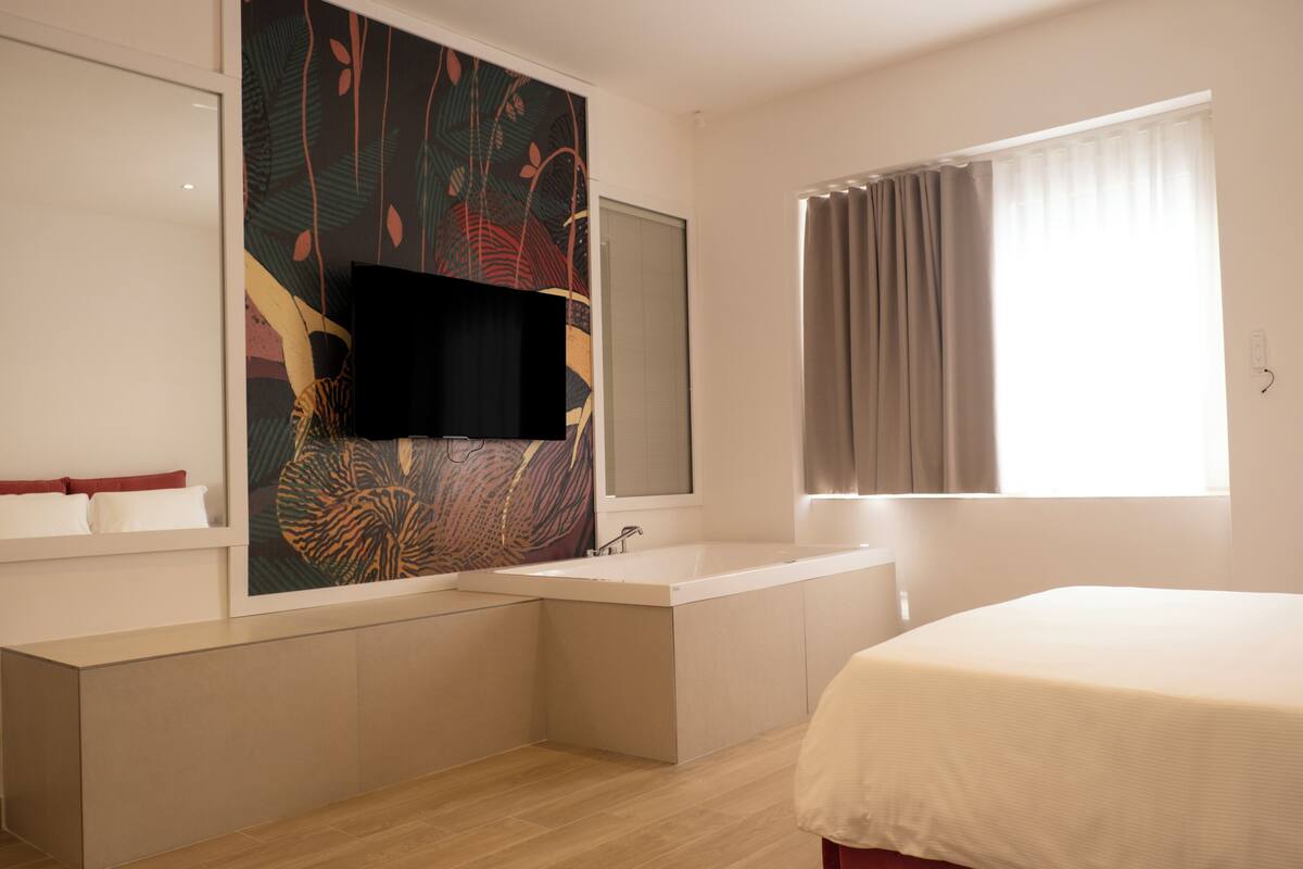 A modern hotel room with a large bed, a built-in bathtub, a wall-mounted TV, and a window with curtains.