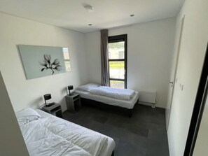 A modern bedroom with two beds, a small bedside table with a lamp, a window with curtains, and a wall-mounted artwork. - Sustainable Home Near the Golf Course (Velsen-Zuid)