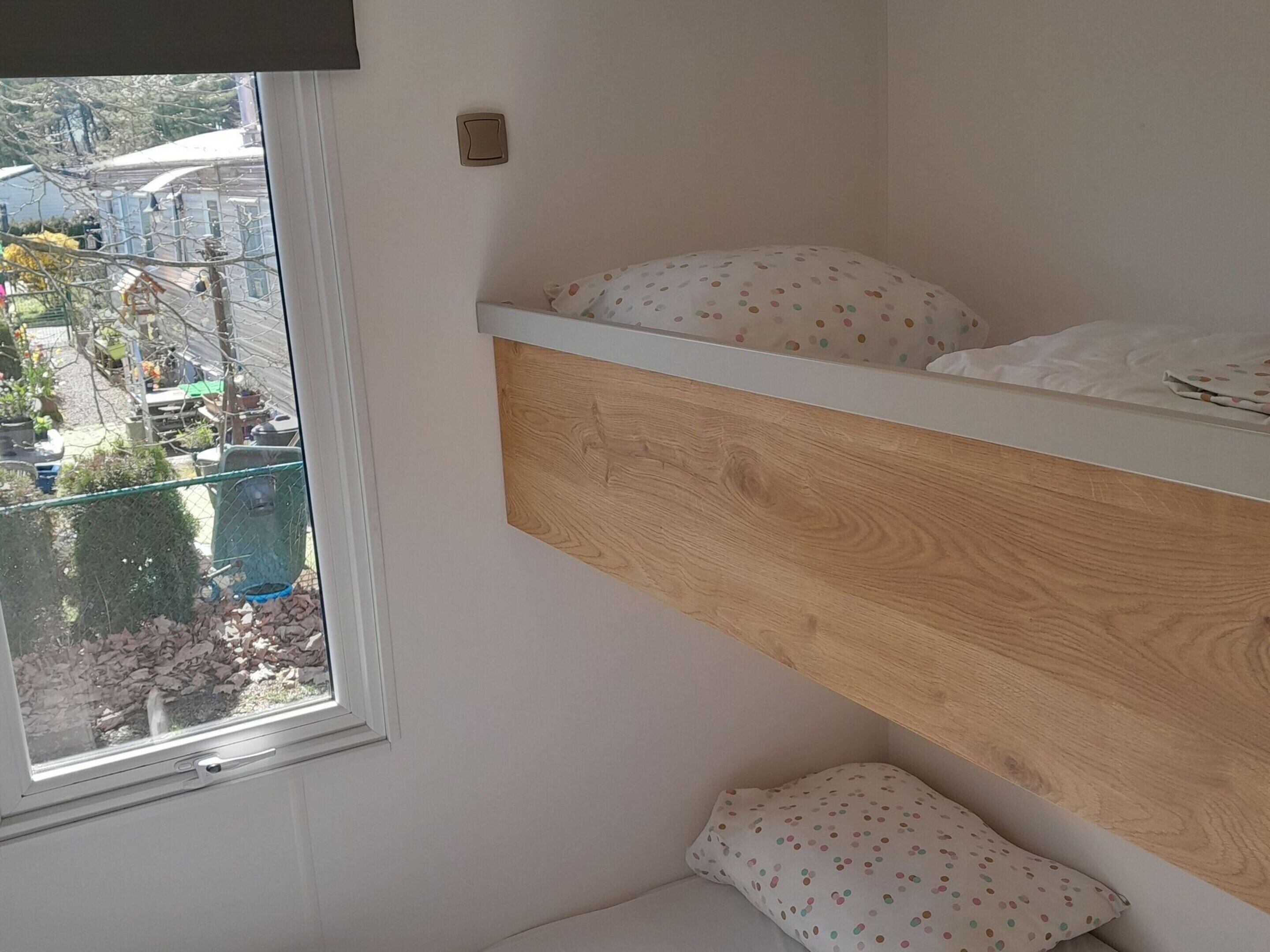 A bunk bed with a wooden headboard, a window with a view of outdoor furniture and plants, and a small bedside table.